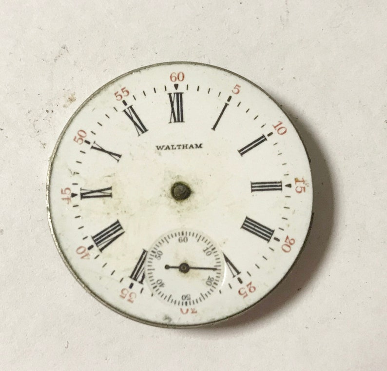 Op de afbeelding: Een wit zakhorloge-horloge met zwarte Romeinse cijfers als uurmarkeringen en zwarte wijzers. De wijzerplaat heeft een kleinere, concentrische cirkel met zwarte markeringen voor seconden. De wijzerplaat is met "WALTHAM" in zwarte letters gemarkeerd.