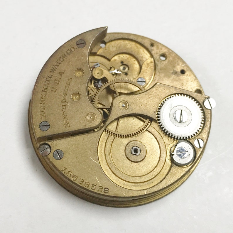 K&ouml;nnte beinhalten: Nahaufnahme eines goldfarbenen Taschenuhrwerks mit der Aufschrift "Elgin National Watch Co. U.S.A. Seven Jewels 19633538".