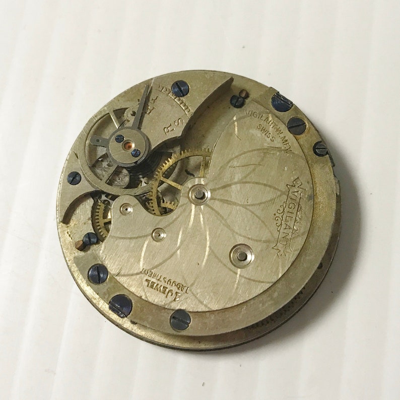 Op de afbeelding: Close-up van de achterkant van een zilveren zakhorloge met een 15-juwelen uurwerk. De wijzerplaat van het horloge heeft een bloemmotief en de tekst "Vigilant Watch Co. Swiss Made" is op de wijzerplaat gedrukt.
