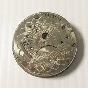May include: Close-up of a silver pocket watch movement with a decorative design and the text "SAFETY PINION" and "ELGIN NAT'L WATCH CO. USA".