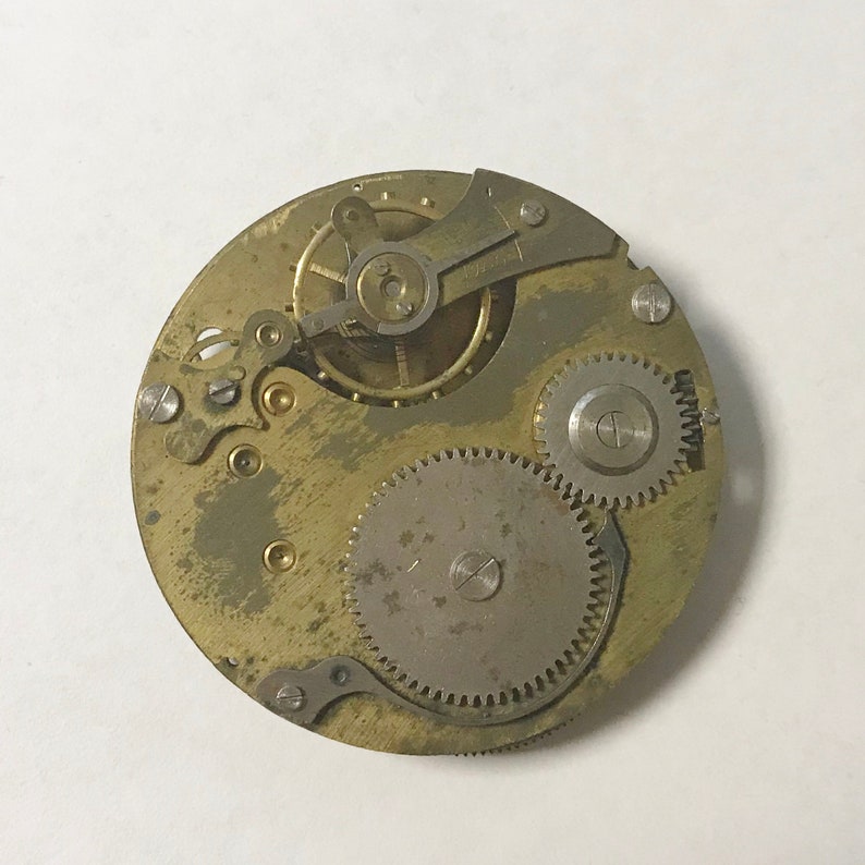 Puede incluir: Primer plano de un movimiento de reloj de tono dorado con engranajes y palancas intrincados. El movimiento est&aacute; expuesto y muestra el funcionamiento interno del reloj.