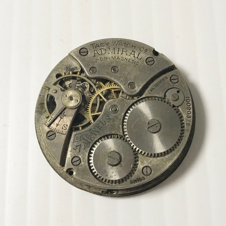 May include: Close-up of a silver pocket watch movement with the text "Tacy Watch Co. Admiral Non-Magnetic" and "7 Jewels" visible. The watch has a 2 adjustment system and the serial number 10090378 is visible.