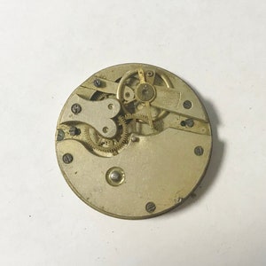 Pu&ograve; includere: Primo piano di un movimento di orologio vintage. Il meccanismo circolare presenta numerosi piccoli ingranaggi, viti e un bilanciere. I componenti metallici hanno un aspetto ossidato e invecchiato, suggerendo l'antichit&agrave; dell'orologio. Il design &egrave; intricato e dettagliato.