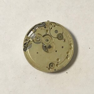 May include: A close-up of a gold-colored watch movement with intricate gears and a silver-colored lever. The movement is labeled "ST. P. 17".