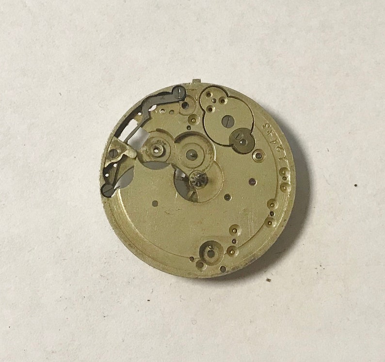 May include: A close-up of a gold-colored watch movement with intricate gears and a silver-colored lever. The movement is labeled "ST. P. 17".