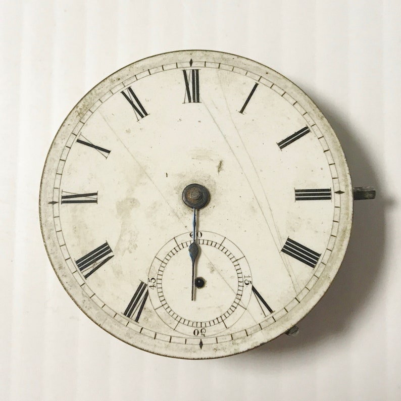May include: A white pocket watch with Roman numeral hour markers and a black second hand. The watch face is slightly aged and has a few scratches.