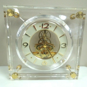 May include: A clear acrylic clock with a gold-toned face and exposed gears. The clock has a white background with black Roman numeral hour markers and black hands.