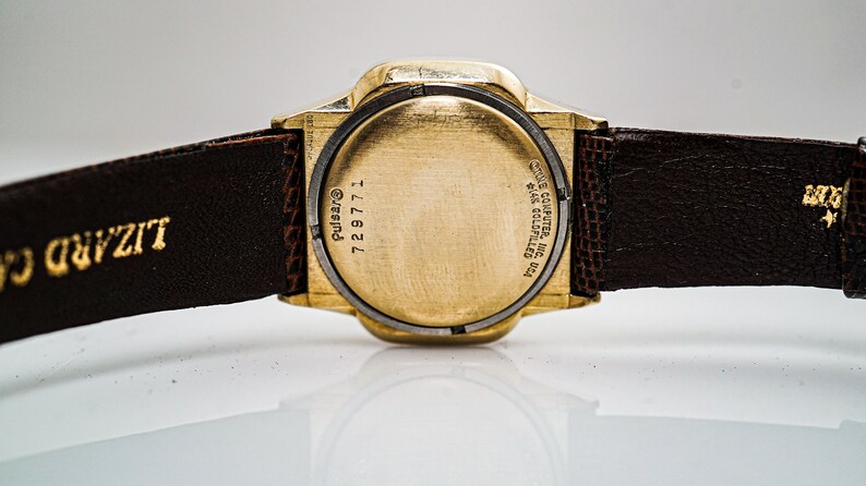 May include: A gold-tone Pulsar watch with a brown leather strap. The back of the watch has the text "Pulsar", "729771", and "ONE COMPUTER INC.  THE COLLECTIBLES  USA". The strap is labelled "Lizard"