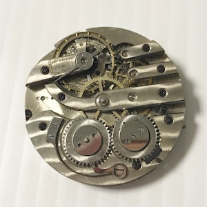 May include: Close-up of a silver pocket watch movement with intricate gears and levers. The watch face is not visible.