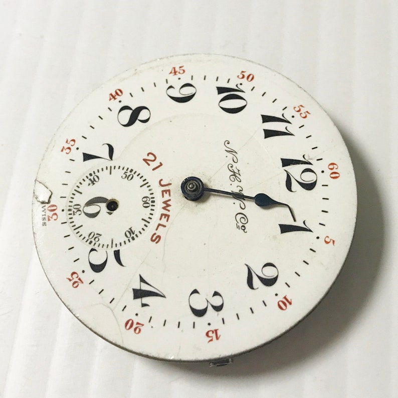 May include: A white pocket watch face with black Roman numeral hour markers and black minute markers. The watch face has a black hand and a small black second hand. The watch face also has the text "21 JEWELS" and "M.H.H. Co.".