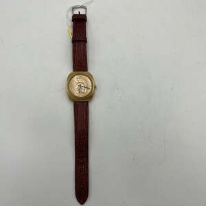Puede incluir: Un reloj de pulsera dorado con una correa de cuero marr&oacute;n. La esfera del reloj presenta un dise&ntilde;o de n&uacute;meros romanos y una peque&ntilde;a imagen de un casco de guerrero.
