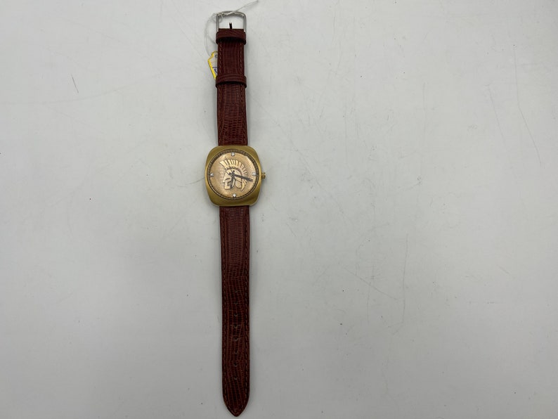 Puede incluir: Un reloj de pulsera dorado con una correa de cuero marr&oacute;n. La esfera del reloj presenta un dise&ntilde;o de n&uacute;meros romanos y una peque&ntilde;a imagen de un casco de guerrero.