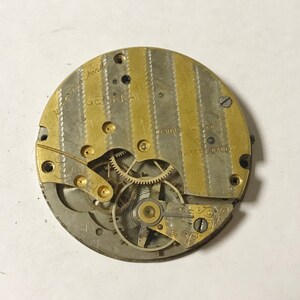 Pode incluir: Close-up de um movimento de rel&oacute;gio de bolso dourado e prateado com o texto "General" e "Double Roller" vis&iacute;vel. O movimento tem uma s&eacute;rie de engrenagens e um design decorativo.