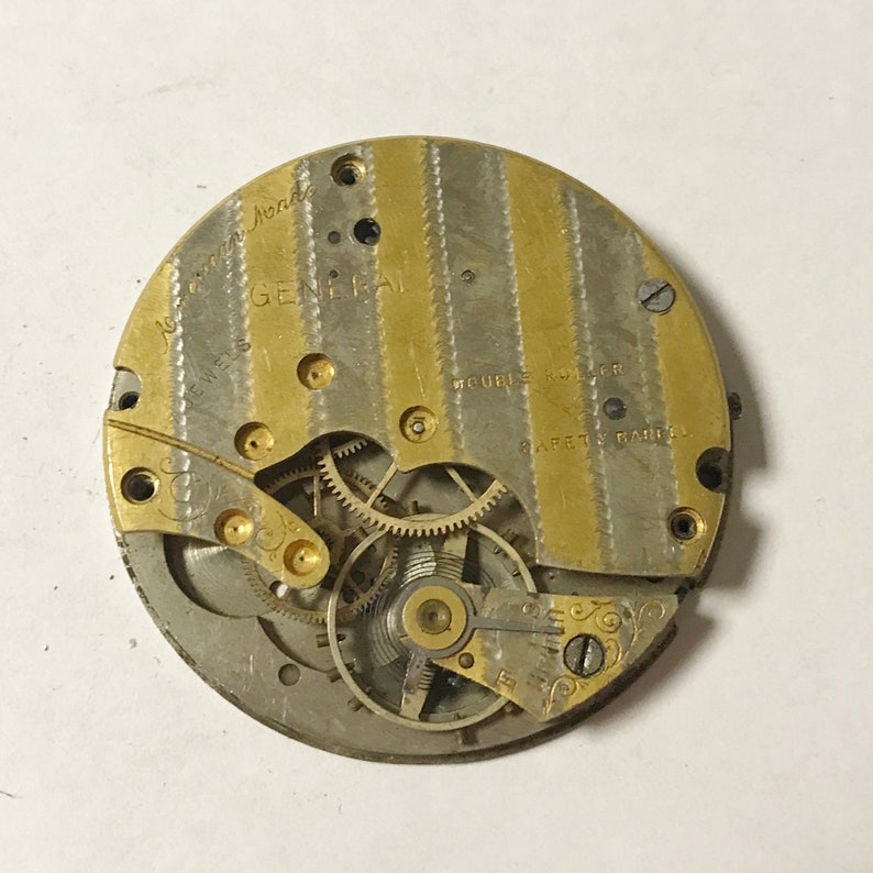Pode incluir: Close-up de um movimento de rel&oacute;gio de bolso dourado e prateado com o texto "General" e "Double Roller" vis&iacute;vel. O movimento tem uma s&eacute;rie de engrenagens e um design decorativo.