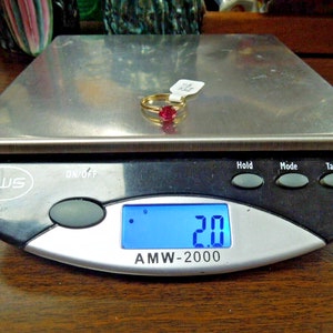 May include: A gold ring with a red gemstone on a digital scale that reads 2.0 grams. The scale has a black base and a silver top.