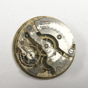 Puede incluir: Primer plano de un movimiento de reloj vintage, mostrando engranajes y componentes intrincados. El mecanismo del reloj tiene el texto "7 JEWELS 1 ADJUSTMENT ENNEFTE SWISS". Las piezas del reloj son de color plateado y dorado.