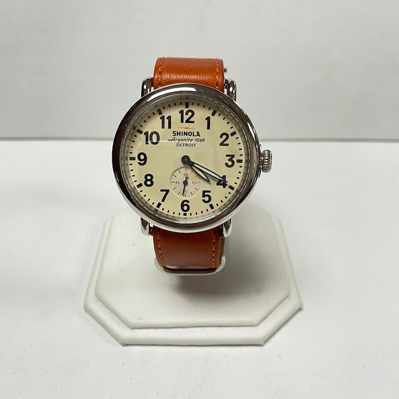May include: A Shinola Argonite-1069 Detroit watch with a cream-colored face, black numerals, and a brown leather band. The watch has a silver-colored case and a small seconds dial. The watch is displayed on a white stand.