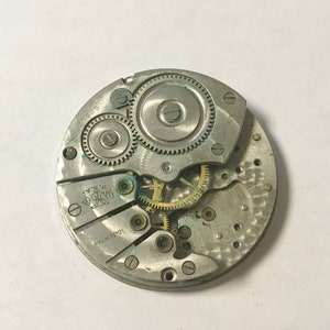 May include: A silver-toned watch movement with intricate gears and a 15 jewel inscription. The movement is in a circular shape and has a diameter of approximately 2.5 inches.