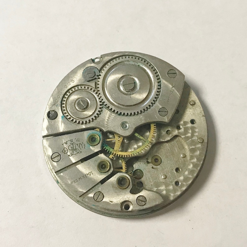 May include: A silver-toned watch movement with intricate gears and a 15 jewel inscription. The movement is in a circular shape and has a diameter of approximately 2.5 inches.