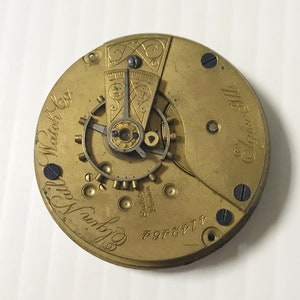 May include: Close-up of the back of a gold-toned pocket watch with the inscription "Elgin National Watch Co." and "Elgin Ill."