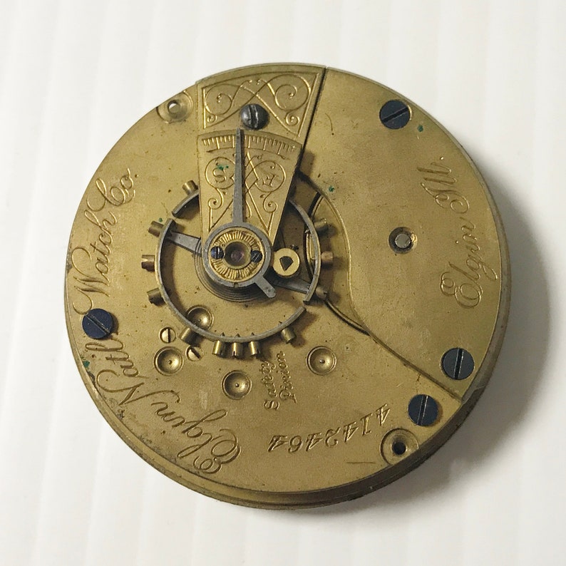 May include: Close-up of the back of a gold-toned pocket watch with the inscription "Elgin National Watch Co." and "Elgin Ill."
