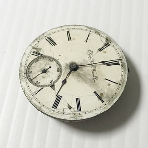 May include: A white pocket watch face with black Roman numeral hour markers and black hands. The watch face is cracked and has a small, round, white sub-dial with black markings. The watch face is inscribed with "Elgin National Watch Co."