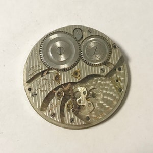 Puede incluir: Un movimiento de reloj de bolsillo plateado con el texto "Adjusted to 3 Positions 1002359" y "Illinois Springfield Watch Co. 19 Jewels" visible. El movimiento tiene una superficie decorativa texturizada.