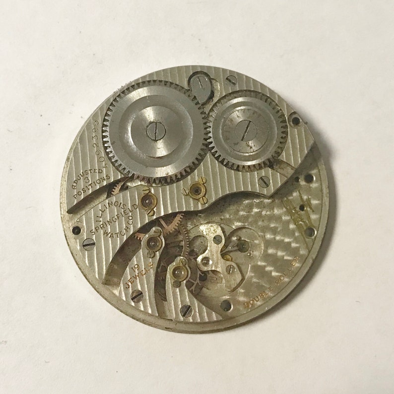 Puede incluir: Un movimiento de reloj de bolsillo plateado con el texto "Adjusted to 3 Positions 1002359" y "Illinois Springfield Watch Co. 19 Jewels" visible. El movimiento tiene una superficie decorativa texturizada.