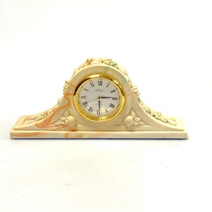 May include: A decorative mantel clock with a cream-colored base and a gold-tone clock face. The clock features Roman numerals and the brand name "Ainsley". The clock is adorned with sculpted fruit and floral details.