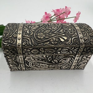May include: A silver metal trinket box with an ornate floral design. The box is closed and has a hinged lid.