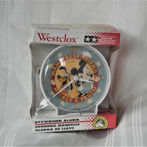 May include: A white key-wound alarm clock with a blue face featuring Mickey Mouse and Pluto. The clock face reads "Man's Best Friend Mickey & Pluto".