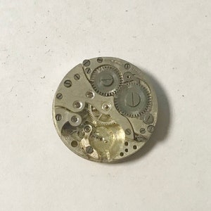 Puede incluir: Primer plano de un movimiento de reloj plateado con engranajes y muelles. El movimiento est&aacute; en una carcasa circular con un di&aacute;metro de aproximadamente 3,8 cm.