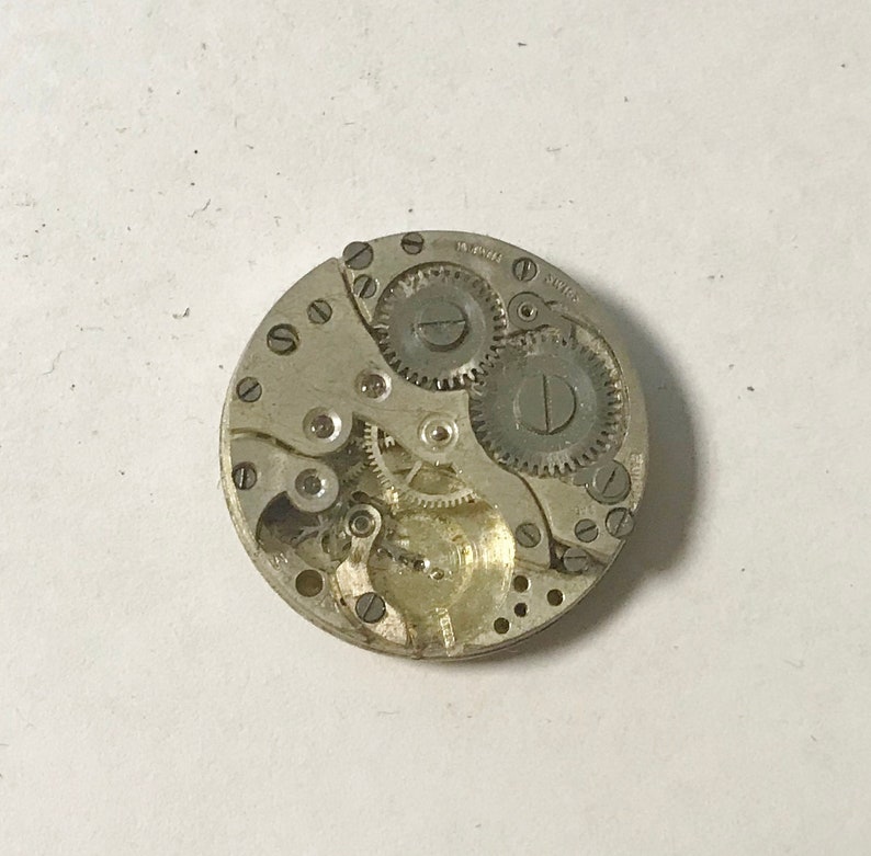 Puede incluir: Primer plano de un movimiento de reloj plateado con engranajes y muelles. El movimiento est&aacute; en una carcasa circular con un di&aacute;metro de aproximadamente 3,8 cm.