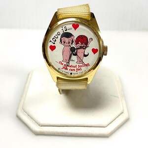 May include: A vintage gold-toned wristwatch with a cream-colored band. The watch face features a cartoon couple with red hearts and the text "love is... the greatest feeling you can feel."