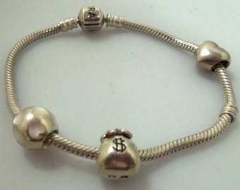 May include: A silver charm bracelet with four charms: a heart, a money bag with a dollar sign, a round charm with white dots, and a charm with the word "Pandora" on it.