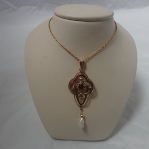 May include: A gold necklace with a pendant featuring a red gemstone and a pearl drop. The pendant is in a quatrefoil shape with intricate details.