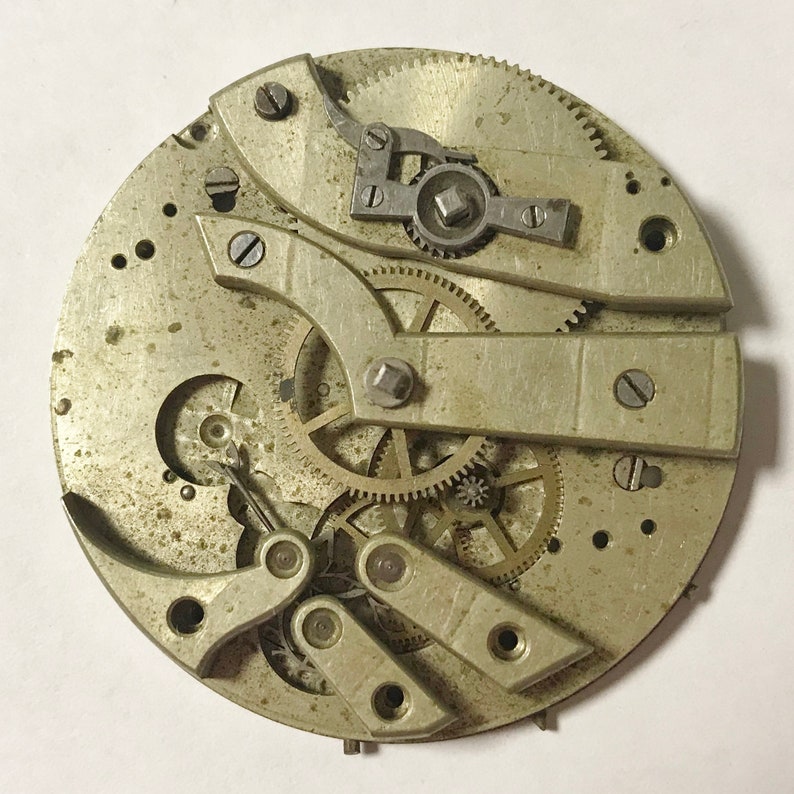 Pu&ograve; includere: Una vista ravvicinata di un movimento di orologio da tasca argentato con ingranaggi e leve intricate. L'orologio &egrave; smontato e disposto su una superficie bianca.