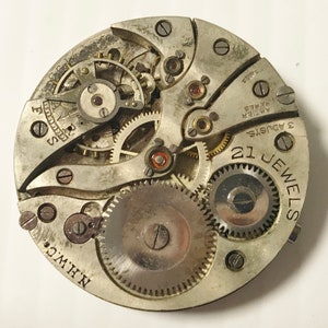 May include: Close-up of a silver watch movement with 21 jewels and 3 adjustments. The movement is labeled "Swiss" and "F.C.C. 17 Jewels." The movement is also labeled "N.H.W.C."