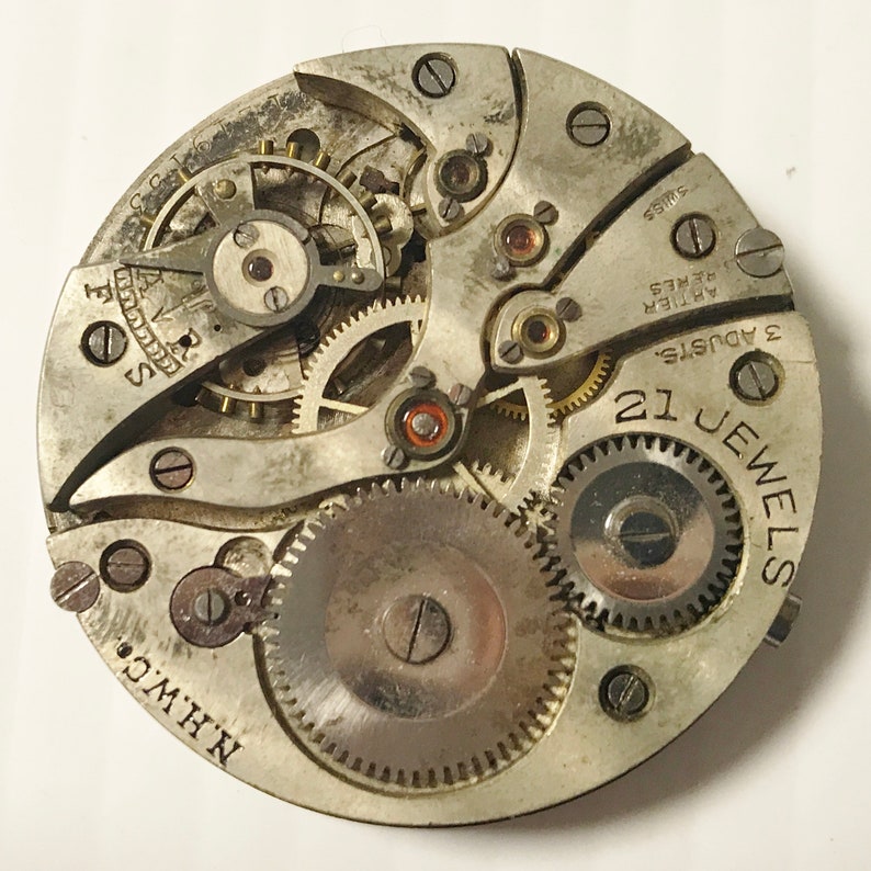 May include: Close-up of a silver watch movement with 21 jewels and 3 adjustments. The movement is labeled "Swiss" and "F.C.C. 17 Jewels." The movement is also labeled "N.H.W.C."