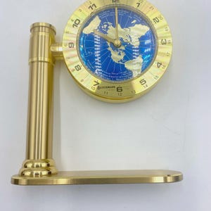 May include: A gold-colored desk clock with a world map design. The clock face is round with a blue background and gold continents. The clock has a gold base and a vertical support. The word "BENCHMARK" is visible.