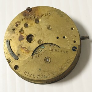 May include: A vintage brass watch movement with visible gears and components. The watch face is inscribed with "W. WELLERY" at the top and "WALTHAM MASS" at the bottom. The serial number "1,682,472" is also visible. The overall color is a warm, aged brass.