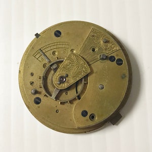 Op de afbeelding: Close-up van een goudkleurig zakhorloge-uurwerk met ingewikkelde gravures en een kleine, ronde wijzerplaat. De wijzerplaat van het horloge is zichtbaar, maar de wijzers ontbreken.