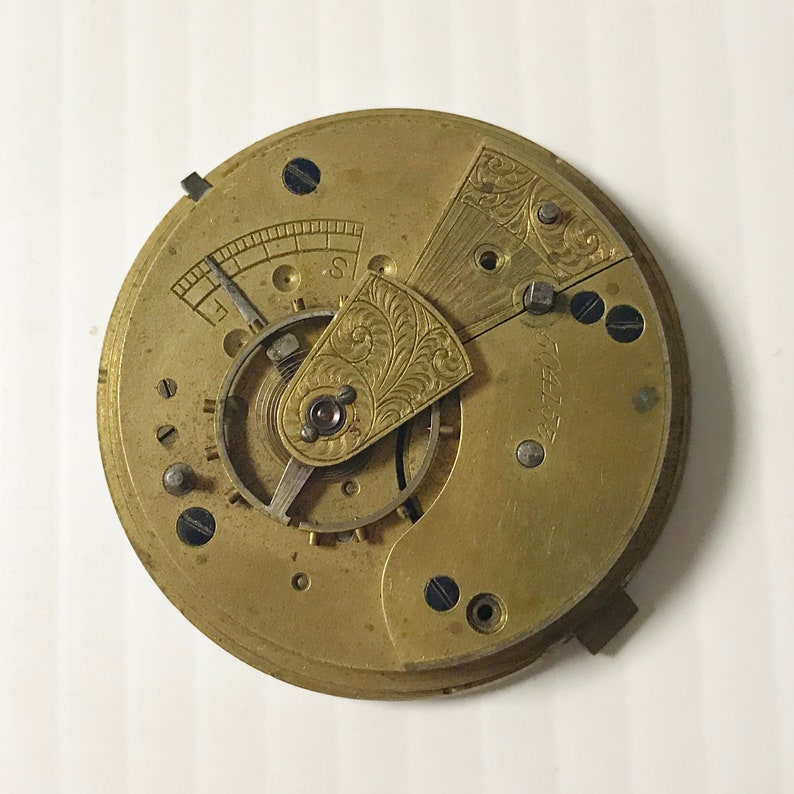 Op de afbeelding: Close-up van een goudkleurig zakhorloge-uurwerk met ingewikkelde gravures en een kleine, ronde wijzerplaat. De wijzerplaat van het horloge is zichtbaar, maar de wijzers ontbreken.