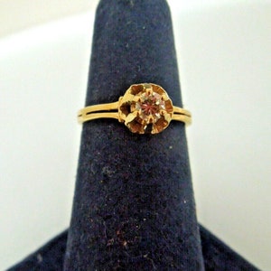 May include: A gold ring with a single round diamond set in a decorative, ornate setting. The ring is on a blue ring sizing mandrel.