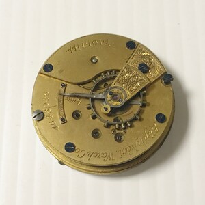 May include: A gold-colored pocket watch movement with the text "Elgin Natl. Watch Co." and "4648036" visible. The movement has a decorative, engraved design on the top.