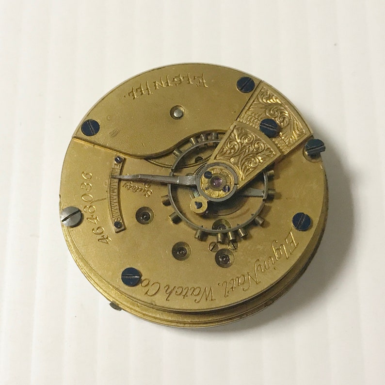 May include: A gold-colored pocket watch movement with the text "Elgin Natl. Watch Co." and "4648036" visible. The movement has a decorative, engraved design on the top.