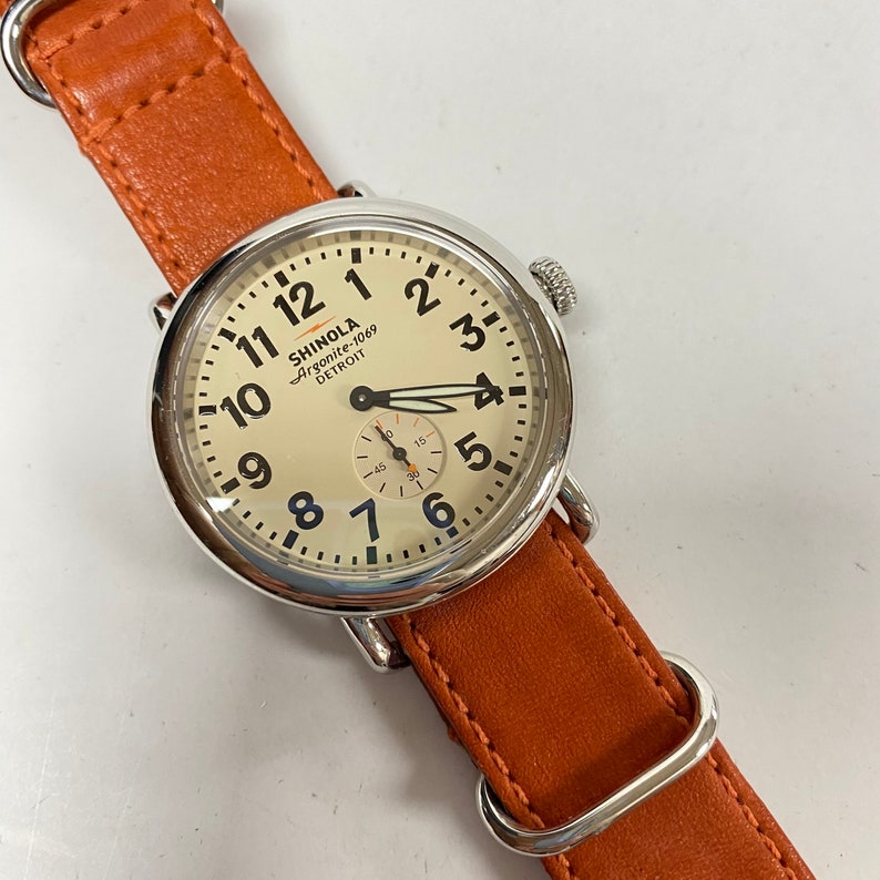 May include: A Shinola Argonite-1069 Detroit watch with a cream-colored face, black numerals, and a small seconds dial. The watch has a stainless steel case and a brown leather strap.