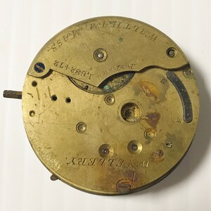 May include: A vintage brass watch movement with the text "WALTHAM, MASS" and "W. ELLERY" engraved on the surface. The movement also features the serial number "1.682.473". The watch mechanism has various gears, screws, and other components.