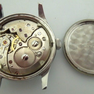 May include: A silver wristwatch with a round face and a silver band. The watch is open, revealing the inner workings of the movement. The back of the watch is also shown, with a smooth, silver surface.