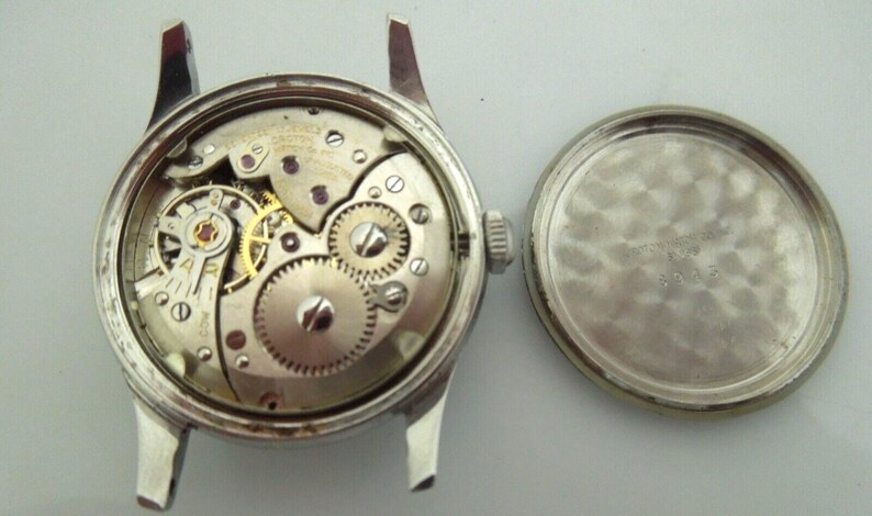 May include: A silver wristwatch with a round face and a silver band. The watch is open, revealing the inner workings of the movement. The back of the watch is also shown, with a smooth, silver surface.
